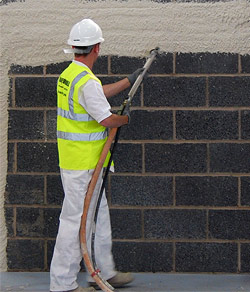 Pracownik w kasku i kamizelce odblaskowej nanosi maszynowo tynk cementowo-wapienny na ścianę z szarych bloczków, widoczny wąż doprowadzający zaprawę.